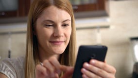 Beautiful young woman with a phablet sitting in kitchen. Browsing Internet and smiling. stock video Beautiful young woman with a phablet sitting in kitchen. Browsing Internet and smiling. stock video