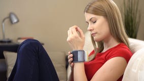 Beautiful young woman sitting on sofa with a wrist tonometer and checking her blood pressure stock video footage Beautiful young woman sitting on sofa with a wrist tonometer and checking her blood pressure stock video footage