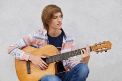 Begåvad tonårs- pojke med skjortan och jeans för moderiktig frisyr som den bärande rymmer den akustiska gitarren som spelar hans arkivbild Begåvad tonårs- pojke med skjortan och jeans för moderiktig frisyr som den bärande rymmer den akustiska gitarren som spelar hans arkivbild