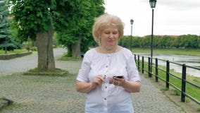 Bejaardegangen langs de waterkant en het gebruiken van smartphone met oortelefoons Rivier op de achtergrond stock video Bejaardegangen langs de waterkant en het gebruiken van smartphone met oortelefoons Rivier op de achtergrond stock video