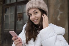 Bella ragazza europea in un rivestimento bianco ed in un cappello tricottato che ascolta la musica con le cuffie che cammina into immagini stock Bella ragazza europea in un rivestimento bianco ed in un cappello tricottato che ascolta la musica con le cuffie che cammina into immagini stock