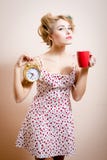 Belle fille de pin-up drôle blonde avec des bigoudis jugeant le réveil et la tasse d'or de la boisson chaude regardant le portrai Image stock Belle fille de pin-up drôle blonde avec des bigoudis jugeant le réveil et la tasse d'or de la boisson chaude regardant le portrai Image stock