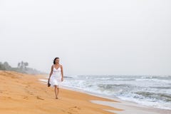 Belle jeune femme dans la robe blanche marchant sur la plage sablonneuse, tenant des sandales Concept de voyage et d'?t? photo libre de droits Belle jeune femme dans la robe blanche marchant sur la plage sablonneuse, tenant des sandales Concept de voyage et d'?t? photo libre de droits