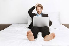 Bello uomo d'affari unshaved biondo che si trova a letto, lavorando al computer portatile, tenente mano sulla testa con colpito fotografia stock Bello uomo d'affari unshaved biondo che si trova a letto, lavorando al computer portatile, tenente mano sulla testa con colpito fotografia stock