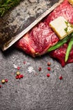 Bifteck de viande crue avec du beurre, les assaisonnements frais et la lame du vieux couteau sur le fond en pierre foncé Photos libres de droits Bifteck de viande crue avec du beurre, les assaisonnements frais et la lame du vieux couteau sur le fond en pierre foncé Photos libres de droits