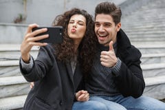 Bild des aufgeregten Paarmannes und der Frau 20s in der warmen Kleidung, selfie Foto am Handy beim Sitzen machend auf der Treppe lizenzfreie stockfotos Bild des aufgeregten Paarmannes und der Frau 20s in der warmen Kleidung, selfie Foto am Handy beim Sitzen machend auf der Treppe lizenzfreie stockfotos