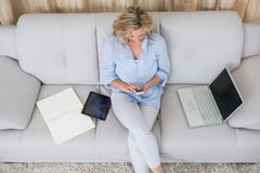 Bionda che si siede sullo strato facendo uso del suo smartphone immagine stock libera da diritti Bionda che si siede sullo strato facendo uso del suo smartphone immagine stock libera da diritti