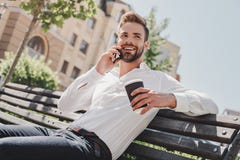 Biznesowa rozmowa Młodego człowieka obsiadanie w parku opowiada na telefonie i pije kawę, zdjęcie stock Biznesowa rozmowa Młodego człowieka obsiadanie w parku opowiada na telefonie i pije kawę, zdjęcie stock