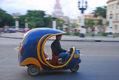 Blue iconic Coco auto rickshaw taxi fast speeding on blurred background of yank tank tanks, Capitol National at Havana, Cuba