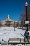 Boston, mA, los E.E.U.U., febrero, 8, 2016: Un hombre joven se encarama en la parte de atr?s de banco al aire libre que lee un li fotografía de archivo Boston, mA, los E.E.U.U., febrero, 8, 2016: Un hombre joven se encarama en la parte de atr?s de banco al aire libre que lee un li fotografía de archivo