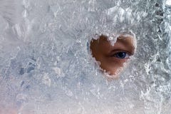 A boy`s blue eye looking through a heart-shaped hole on a frozen glass window