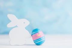 Bright Easter Background: White Wooden Bunny With Paited Egg Against Blue Wall. Holiday Concept Stock Photos