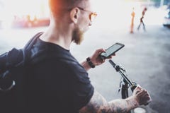 Brodaty mięśniowy tatuujący modniś w okularach przeciwsłonecznych używać smartphone po jechać elektryczną hulajnogą w mieście obraz royalty free Brodaty mięśniowy tatuujący modniś w okularach przeciwsłonecznych używać smartphone po jechać elektryczną hulajnogą w mieście obraz royalty free