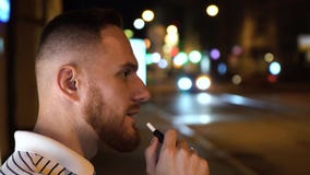 Brodaty mężczyzna w pasiastego t koszula dymu elektronicznych papierosowych iqos przy nocą na ulicie z samochodami na tle zdjęcie wideo Brodaty mężczyzna w pasiastego t koszula dymu elektronicznych papierosowych iqos przy nocą na ulicie z samochodami na tle zdjęcie wideo
