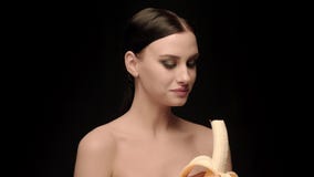 Brunette μόδας που στέκεται στο μαύρο υπόβαθρο τοίχων στο στούντιο, που τρώει μια μπανάνα απόθεμα βίντεο Brunette μόδας που στέκεται στο μαύρο υπόβαθρο τοίχων στο στούντιο, που τρώει μια μπανάνα απόθεμα βίντεο