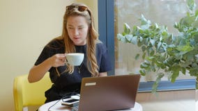 Business woman sitting at a table in a cafe drinking coffee and working on a laptop stock video footage Business woman sitting at a table in a cafe drinking coffee and working on a laptop stock video footage