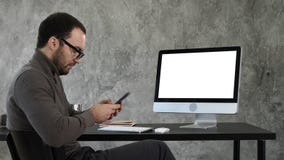 Businessman looking at his smartphone messaging and sitting near computer screen. White Display. stock photography Businessman looking at his smartphone messaging and sitting near computer screen. White Display. stock photography