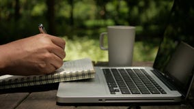 Bärbar dator för bruk för manfreelancer som en märkes- skriver på anteckningsboken och dricker kaffe på trädgården arkivfilmer Bärbar dator för bruk för manfreelancer som en märkes- skriver på anteckningsboken och dricker kaffe på trädgården arkivfilmer