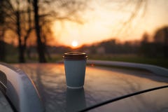 Caff? della tazza di carta al tramonto che sta su un tetto dell'automobile con bello dal bokeh del fuoco immagine stock libera da diritti Caff? della tazza di carta al tramonto che sta su un tetto dell'automobile con bello dal bokeh del fuoco immagine stock libera da diritti