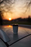 Caff? della tazza di carta al tramonto che sta su un tetto dell'automobile con bello dal bokeh del fuoco fotografia stock Caff? della tazza di carta al tramonto che sta su un tetto dell'automobile con bello dal bokeh del fuoco fotografia stock