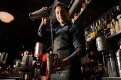 Camarero sonriente joven que vierte la bebida alcohólica a partir de un vidrio del metal en otro imagenes de archivo Camarero sonriente joven que vierte la bebida alcohólica a partir de un vidrio del metal en otro imagenes de archivo