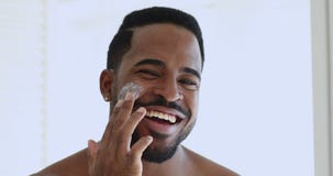 Charming smiling african guy applying facial cream looking at camera