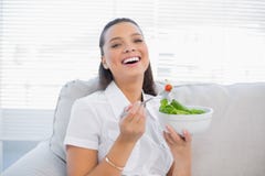 Cheerful pretty woman holding healthy salad sitting on sofa. In bright living room Royalty Free Stock Photography Cheerful pretty woman holding healthy salad sitting on sofa. In bright living room Royalty Free Stock Photography