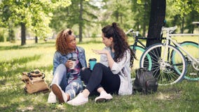 Cheerful young lady is talking to her African American friend and drinking takeout coffee in park on nice green lawn stock video footage Cheerful young lady is talking to her African American friend and drinking takeout coffee in park on nice green lawn stock video footage