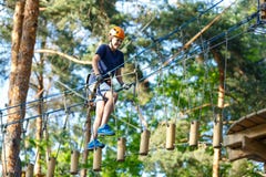 Child In Forest Adventure Park. Kid In Orange Helmet And Blue T Shirt Climbs On High Rope Trail. Stock Photo Child In Forest Adventure Park. Kid In Orange Helmet And Blue T Shirt Climbs On High Rope Trail. Stock Photo