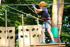 Child In Forest Adventure Park. Kid In Orange Helmet And Blue T Shirt Climbs On High Rope Trail. Agility Skills Royalty Free Stock Images Child In Forest Adventure Park. Kid In Orange Helmet And Blue T Shirt Climbs On High Rope Trail. Agility Skills Royalty Free Stock Images