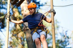 Child In Forest Adventure Park. Kid In Orange Helmet And Blue T Shirt Climbs On High Rope Trail. Agility Skills Stock Image