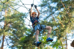 Child In Forest Adventure Park. Kid In Orange Helmet And Blue T Shirt Climbs On High Rope Trail. Agility Skills Stock Photos