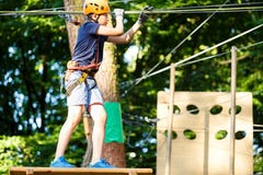 Child In Forest Adventure Park. Kid In Orange Helmet And Blue T Shirt Climbs On High Rope Trail. Agility Skills And Climbing Stock Photo