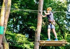 Child In Forest Adventure Park. Kid In Orange Helmet And Blue T Shirt Climbs On High Rope Trail. Agility Skills And Climbing Royalty Free Stock Photo Child In Forest Adventure Park. Kid In Orange Helmet And Blue T Shirt Climbs On High Rope Trail. Agility Skills And Climbing Royalty Free Stock Photo