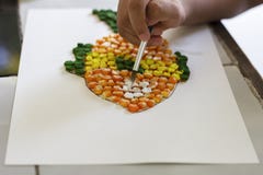 Child`s hands holding a paintbrush coloring corn kernels while doing art fish shape on white paper.
