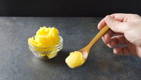 Close up da colher com manteiga carified contra da bacia de vidro completamente de manteiga deliciosa e saudável fresco feito imagem de stock Close up da colher com manteiga carified contra da bacia de vidro completamente de manteiga deliciosa e saudável fresco feito imagem de stock