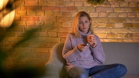 Close-upspruit van volwassen Kaukasische blonde vrouwelijke het letten op TV die een kop van warme thee houden terwijl binnen het stock video Close-upspruit van volwassen Kaukasische blonde vrouwelijke het letten op TV die een kop van warme thee houden terwijl binnen het stock video