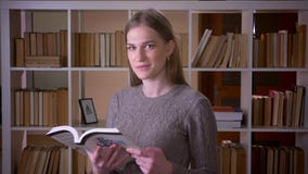 Closeup portrait of young attractive female student reading a book and smiling happily looking at camera in the college stock footage Closeup portrait of young attractive female student reading a book and smiling happily looking at camera in the college stock footage