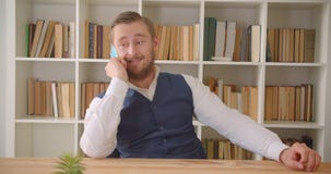 Closeup portrait of young caucasian businessman having a phone call on the workplace indoors with bookshelves on the stock video Closeup portrait of young caucasian businessman having a phone call on the workplace indoors with bookshelves on the stock video