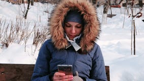 Closeupkvinnan sitter på bänk, och bläddra mobiltelefonen i vinter parkera i staden under dagen i snöig väder stock video Closeupkvinnan sitter på bänk, och bläddra mobiltelefonen i vinter parkera i staden under dagen i snöig väder stock video
