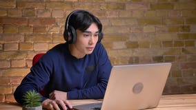 Closeupstående av den unga attraktiva koreanska mannen som använder bärbara datorn och sätter på hans hörlurar stock video Closeupstående av den unga attraktiva koreanska mannen som använder bärbara datorn och sätter på hans hörlurar stock video