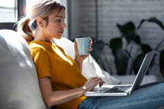 Confident Young Woman Working With Her Laptop While Drinking A Cup Of Coffee Sitting On A Couch At Home Stock Photos