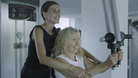Contratan a la mujer mayor en un simulador al gimnasio con un instructor personal la hija ayuda a la mamá en el gimnasio imagen de archivo libre de regalías Contratan a la mujer mayor en un simulador al gimnasio con un instructor personal la hija ayuda a la mamá en el gimnasio imagen de archivo libre de regalías