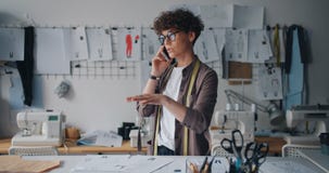 Costureiros fêmeas que falam no telefone celular que olha os esboços que trabalham no estúdio vídeos de arquivo Costureiros fêmeas que falam no telefone celular que olha os esboços que trabalham no estúdio vídeos de arquivo