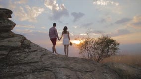 Couple stand on rock and watching sunset on top of the mountain. Girl is having flower in her hand Couple stand on rock and watching sunset on top of the mountain. Girl is having flower in her hand