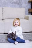 Cute little girl with grey Easter rabbit, bunny. Blonde baby girl smiling in white sweater and blue jeans sitting at home on the c Royalty Free Stock Image Cute little girl with grey Easter rabbit, bunny. Blonde baby girl smiling in white sweater and blue jeans sitting at home on the c Royalty Free Stock Image