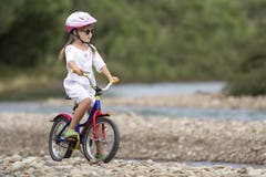Cute young girl in white clothing, sunglasses with long braids wearing pink safety helmet riding child bicycle on pebbled river stock photos Cute young girl in white clothing, sunglasses with long braids wearing pink safety helmet riding child bicycle on pebbled river stock photos