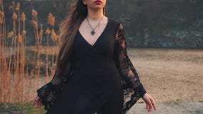 Dark queen on shore of magical lake, evil witch in chic black dress with loose lace sleeves and high leather boots, girl