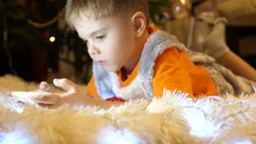 Das Kind liegt auf einer weichen weißen Decke im Kinderzimmer Er passt Karikaturen auf dem Smartphone auf Weihnachten stock video footage Das Kind liegt auf einer weichen weißen Decke im Kinderzimmer Er passt Karikaturen auf dem Smartphone auf Weihnachten stock video footage