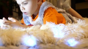Das Kind liegt auf einer weichen weißen Decke im Kinderzimmer Er passt Karikaturen auf dem Smartphone auf Weihnachten stock video Das Kind liegt auf einer weichen weißen Decke im Kinderzimmer Er passt Karikaturen auf dem Smartphone auf Weihnachten stock video